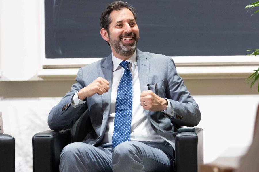 David Folkenflik, with black hair, salt and pepper beard and mustache, in suit and tie, laughing, seated in an armchair.