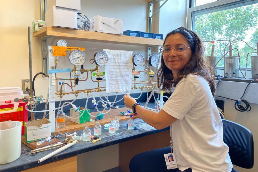 Ligia Coelho, with wire glasses and t-shirt, smiling at the camera next to her lab bench with dials and beakers and wires connecting them