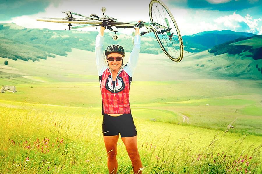 Person wearing a bike helmet and shorts, holds a bicycle over her head. Mountains in the background