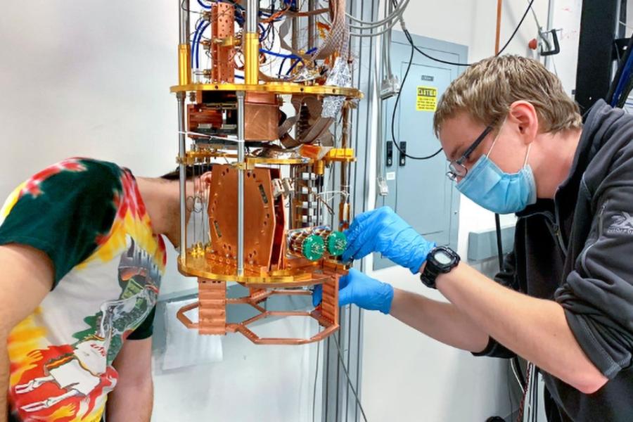 Two people lean toward a complicated scientific instrument featuring gold and orange metal parts; it's about the size of a coffeemaker