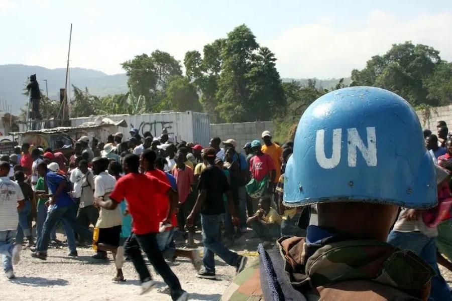 A UN blue peacekeeper's helmet in the foreground; facing a crowd of people
