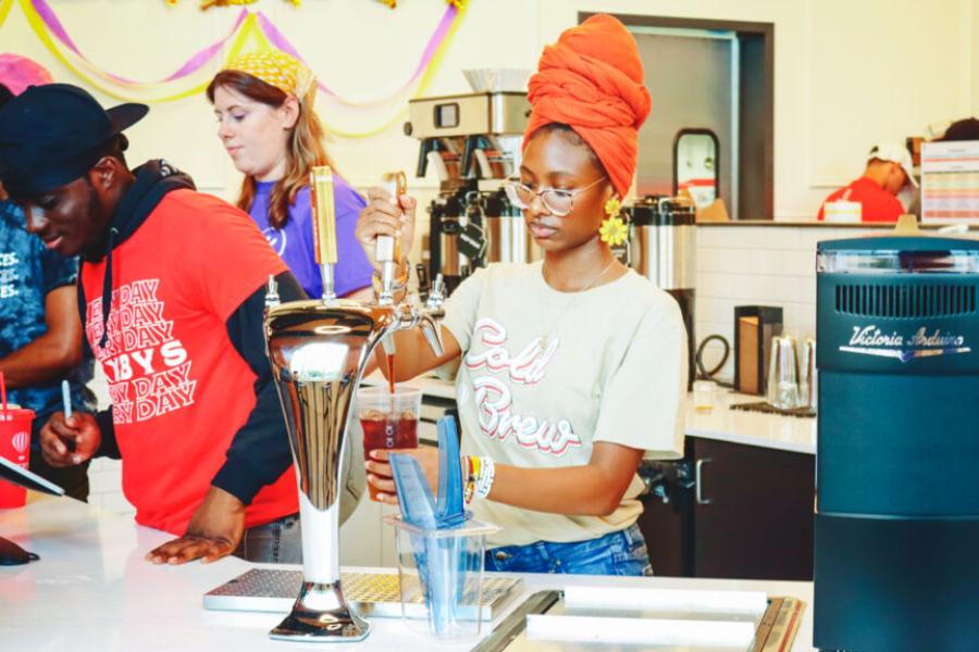 College students with hats and scarves covering their hair and Saxbys t-shirts make coffee.
