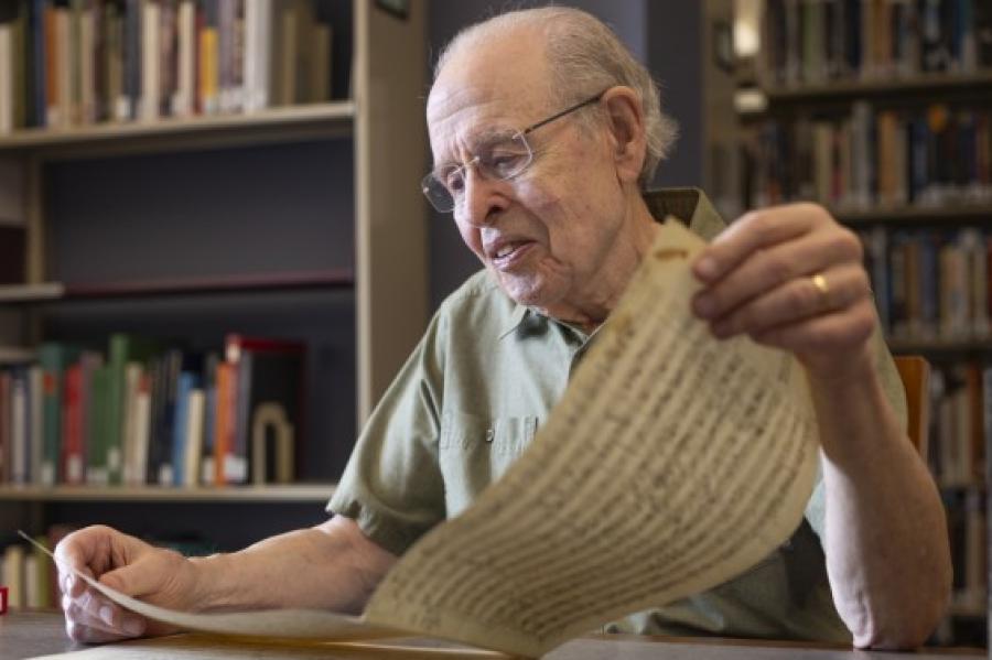 Neal Zaslaw, in glasses and short-sleeved button-down shirt, looking at a musical score long enough that he is holding it in both hands.
