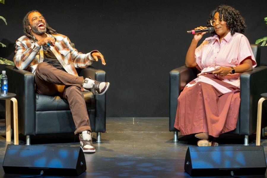 Two people on a stage, sitting in arm chairs, holding microphones