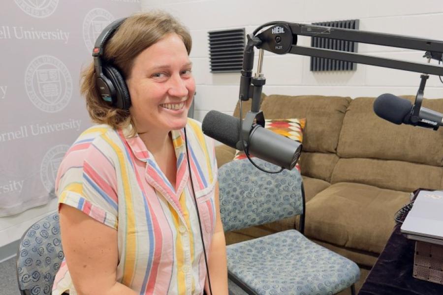 Alexis Boyce, wearing headphones in front of a big microphone, smiles at the camera, wearing a bright colored, striped shirt.