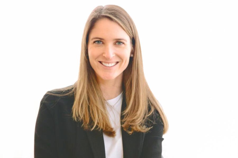 Jenna Barnes, dressed in jeans, open sweater and t-shirt, with long brown hair, seated and smiling.