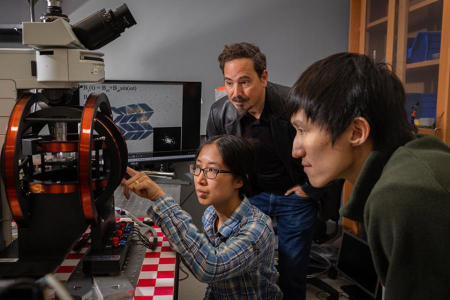 Three people look intensely at a small black and red machine in a science laboratory