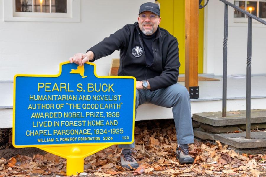 Person sits on a porch with one hand on a blue historic marker