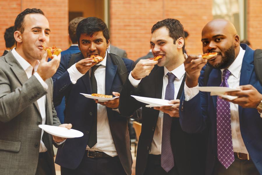 Four people wearing suits, all chomping on slices of pizza and holding plates