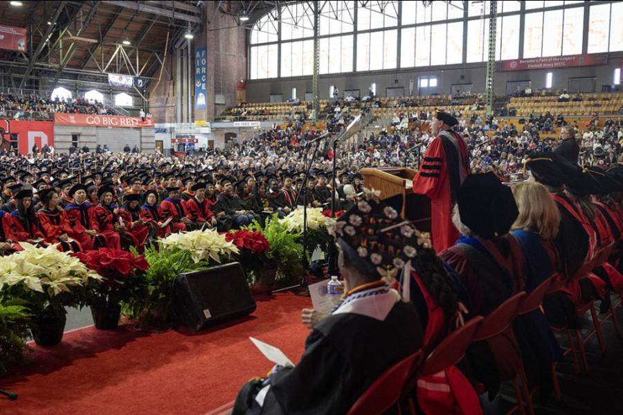 Large indoor gymnasium (Cornell's Barton Hall) decorated with pointsettias and filled with people wearing caps and gowns