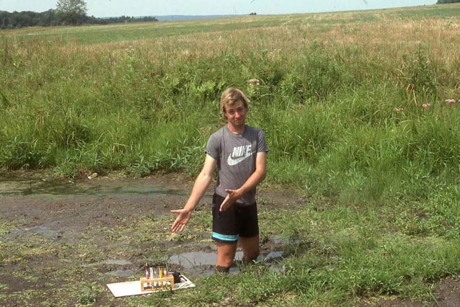 Smiling man in t-shirt and shorts standing in a bog up to his thighs pointing toward a wooden platform in the water.