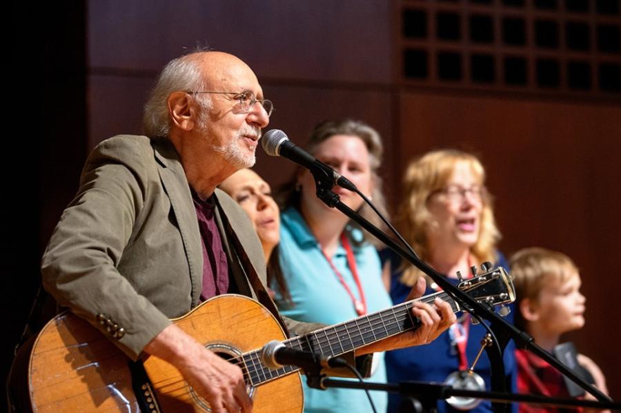 Peter Yarrow with guitar, tan jacket, white hair just at the back of his head, white beard and glasses in front of a microphone with three women and a child standing next to him