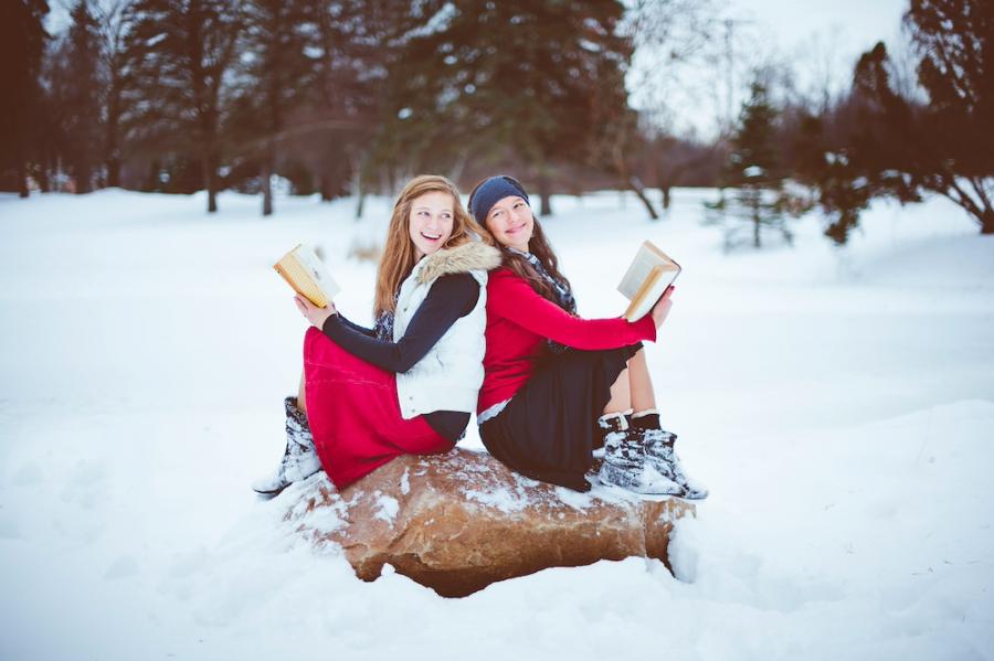 Two people wearing fashionable red, white and black winter clothing sit back to back on a large rock, each holding a book. They are surrounded by snow