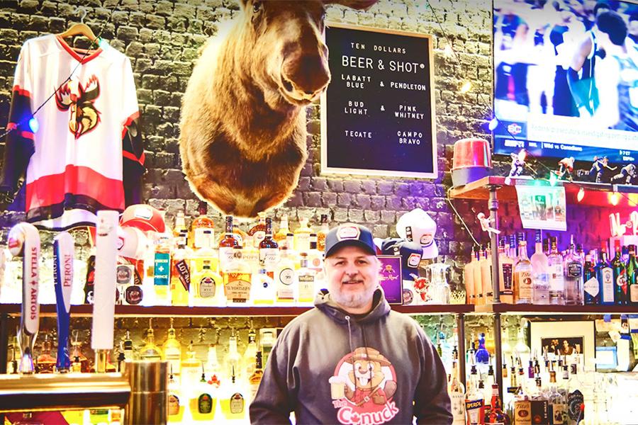 Person standing behind a bar decorated with a hockey jersey, a beer & shot list and a mounted moose head. Lots of neon lights and two illuminated shelves of liquor bottles