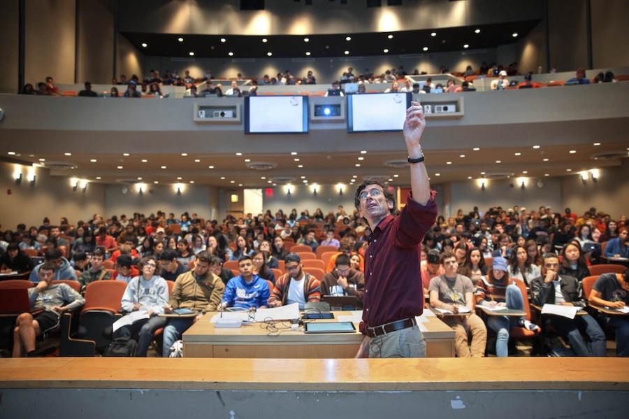 An instructor on a platform, pointing upwards while hundreds of students watch from seats in an auditorium