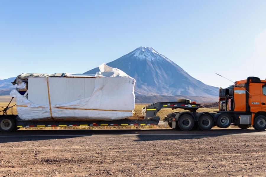 A huge truck pulling an enormous container along a dirt road through a desert, with a snow capped mountain in the distance.