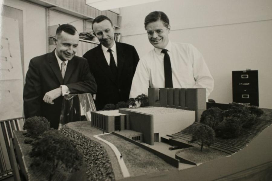 Black and white historic image of three people, wearing ties, looking over a tabletop model of a building