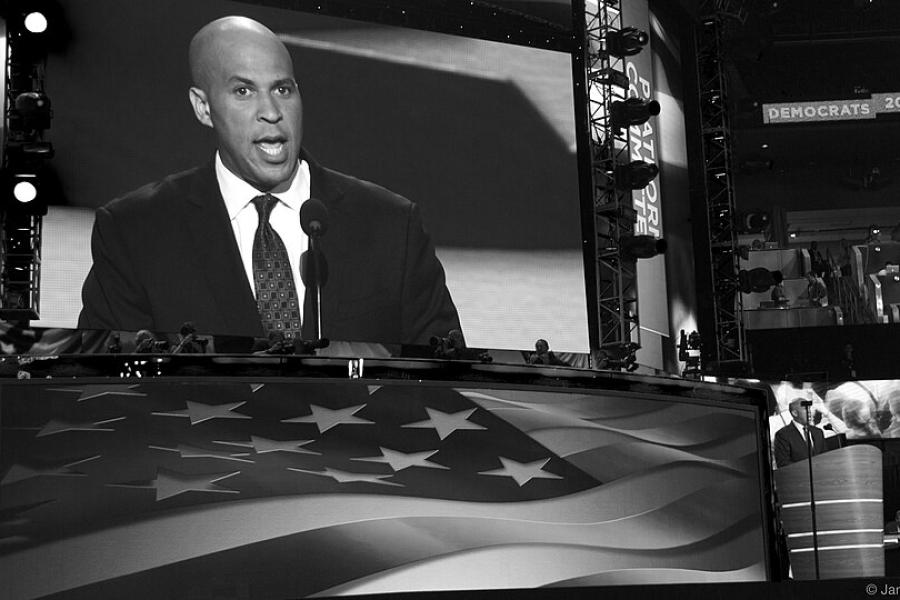 Corey Booker in suit and tie on a large screen with a huge American flag in front of it,