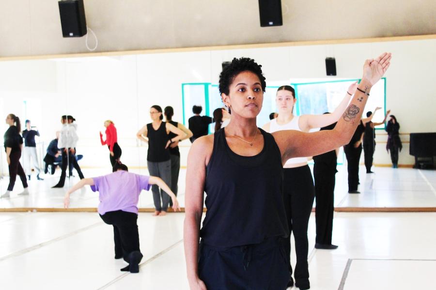 A dancer in the foreground stands upright, holding their left arm at an angle. In the background another dancer replicates the pose, and several other people take different poses or stand around