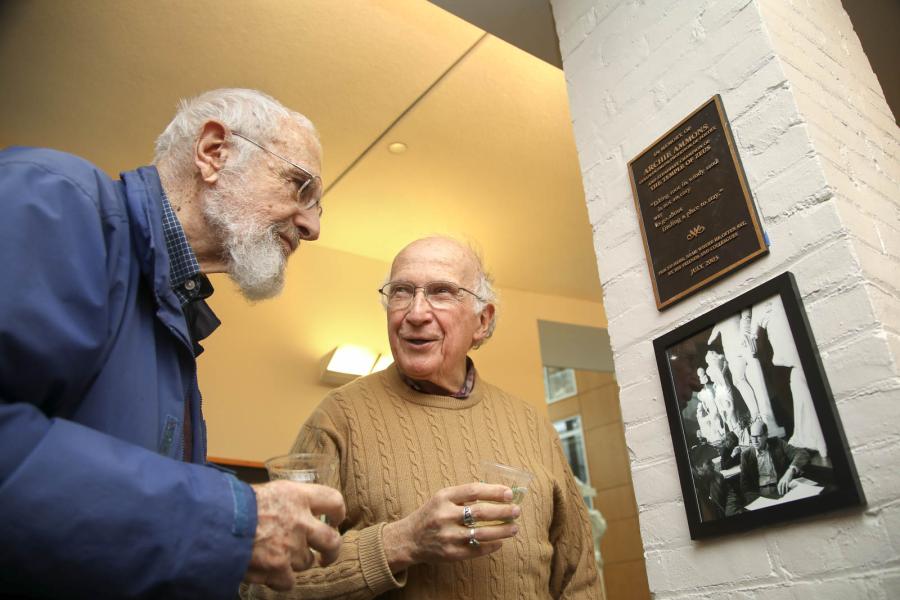 Roald Hoffmann, right, talks about Archie Ammons with another man. A plaque dedicated to Ammons and a photo of him are hung on the column on the right.