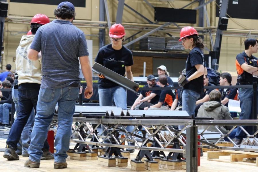 People in red hard hats construct a structure on the floor of a large indoor space