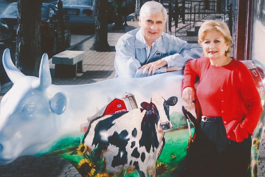 A white haired man and a woman in bright red shirt and sweater stand leaning against a cow with a painting of a cow on it, with a barn in the back and sunflowers and grass on its legs.