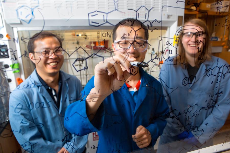 Professors writing on a clear board with two men in the background