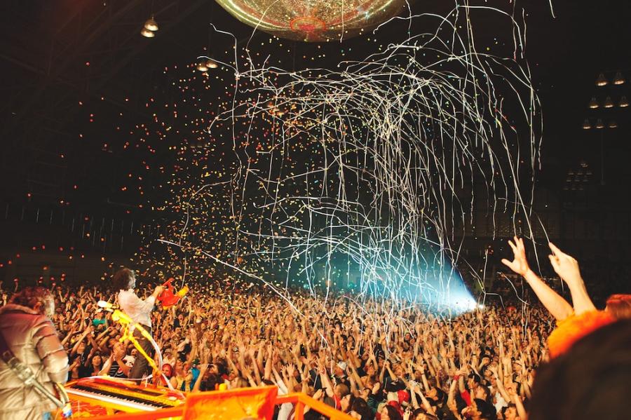 A crowd raises their hands with excitement as a band plays on a stage (barely in the image) and confetti rains down from above