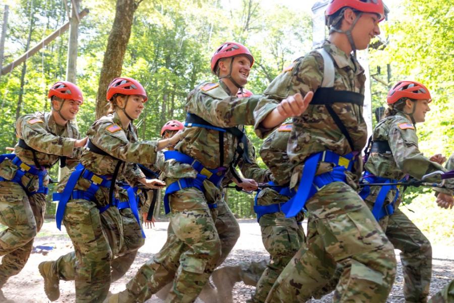Several people in Army fatigues and red helmets run, strapped together