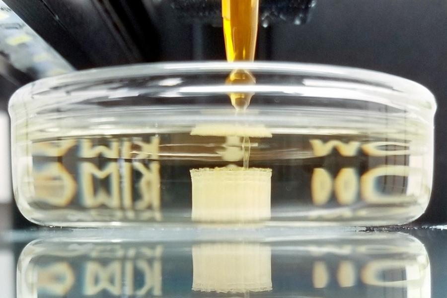 Yellow probiscus descends into a clear dish to deposit  during the 3D printing process, where it self-assembles before being heat-treated into a crystalline superconductor.