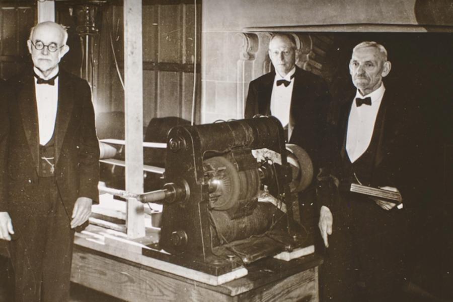 Historic black and white photo of three people in evening coats standing around a machine