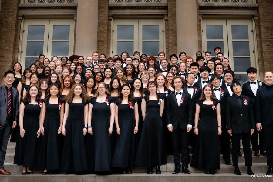 Members of a large choir, wearing black performance outfits, pose as a group on the steps of a building with stone columns