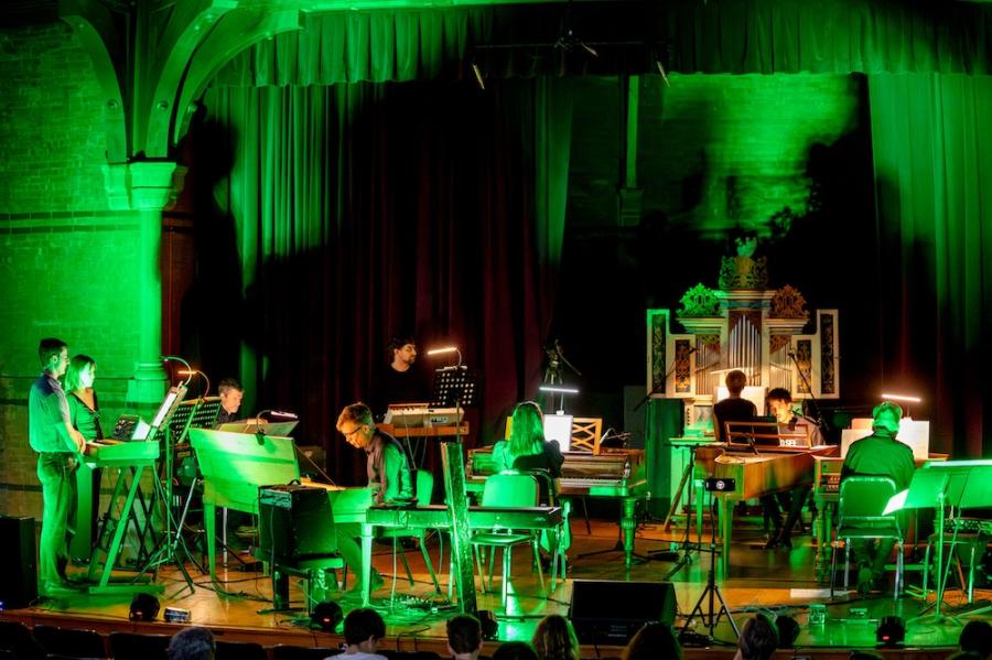 A stage bathed in green light: several people play on keyboard instruments, including standing sythesizers, pianos, and an organ