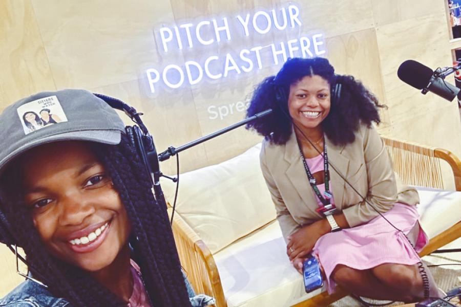 Two people wearing headphones and recording equipment under a neon sign: Pitch your podcast here