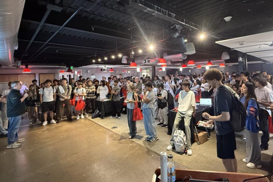Lots of students standing in a big space with bright lights overhead facing someone with a microphone.