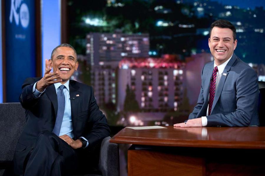 Two people sit at a desk on the set of a late night TV chat show
