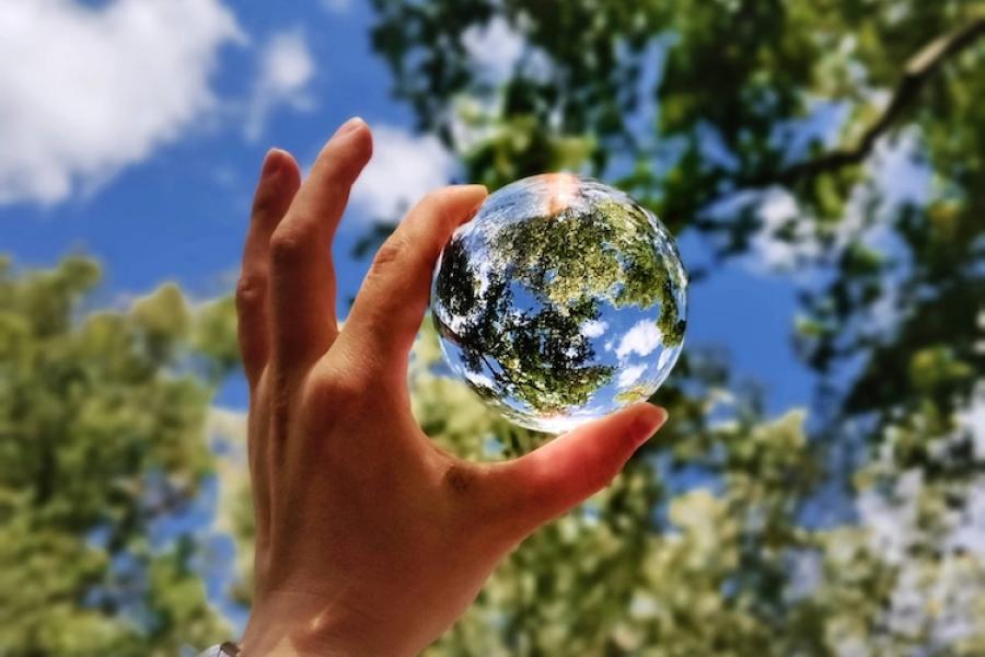 Hand holding a glass sphere up toward the sky so it reflects trees and clouds, making the ball look like a tiny planet Earth