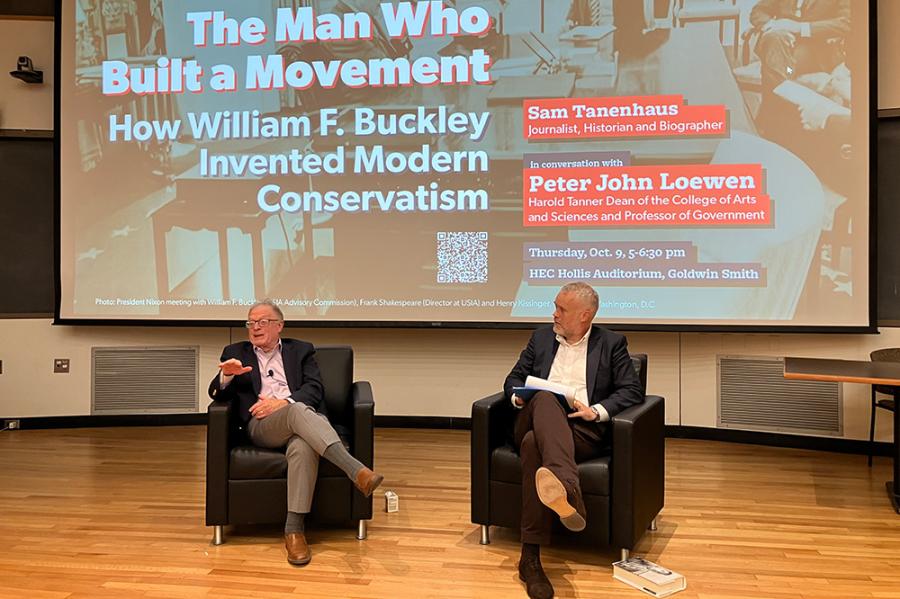 Two people sitting in arm chairs, conversing in front of an audience