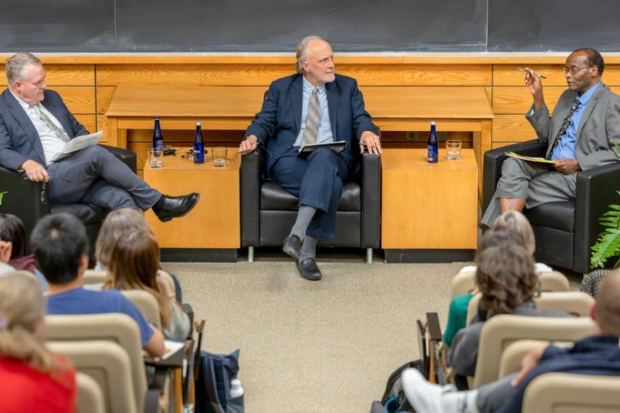 Three people sit in chairs in front of audience during a debate