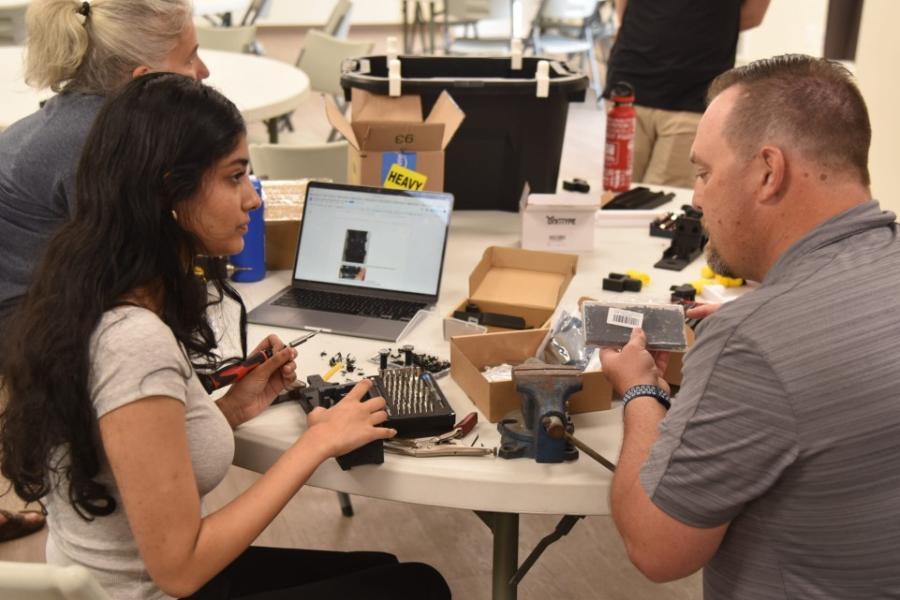 Two people sit at a table loaded with equipment, assembling a robot