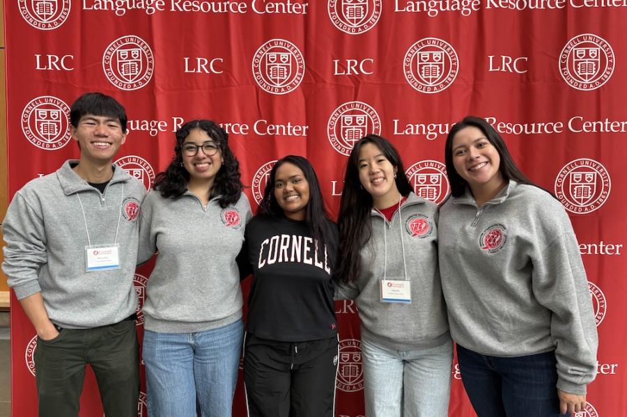 five people wearing Cornell sweatshirts pose in front of a red backdrop
