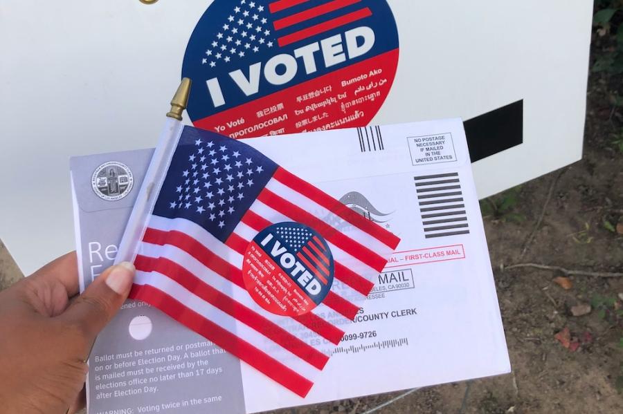 Hand holding a small American flag, a mail-in ballot