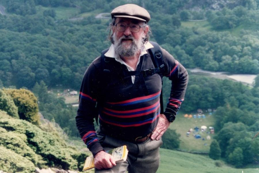 Person wearing a cap, sweater, wool hiking pants and sturdy boots, poses on rocky ground of a very steep hill