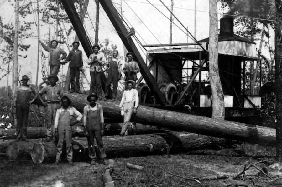 historical black and white photo of about 10 lumber workers posing on a mechanized crane in the woods. They are standing on felled trees.