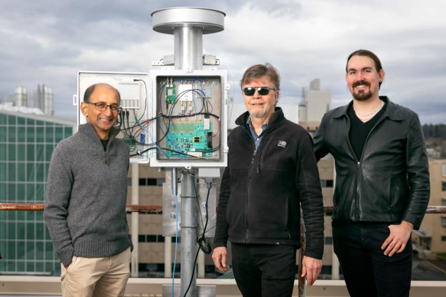 Three people stand next to a silver device, about a foot taller than the people, made of a post, a box with electronics inside, and a circular top