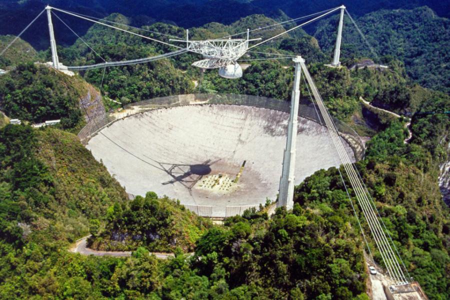 Large concrete dish surrounded by three poles and wires; a mechanism is suspended over the dish. the whole thing, a telescope, is surrounded by lush trees