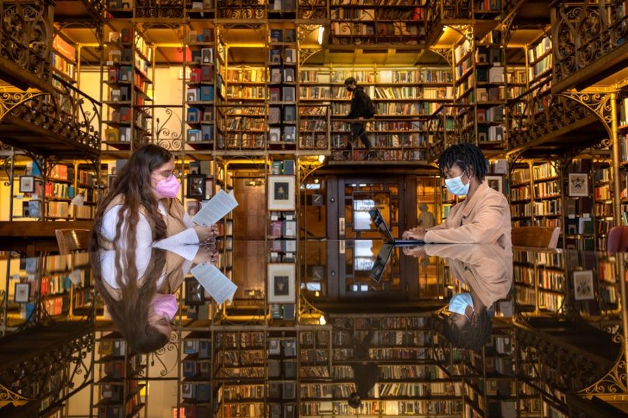 Two people sitting at a table, reading in a room full of books on shelves; the books are reflected on the surface of the table, creating a sense of immersion