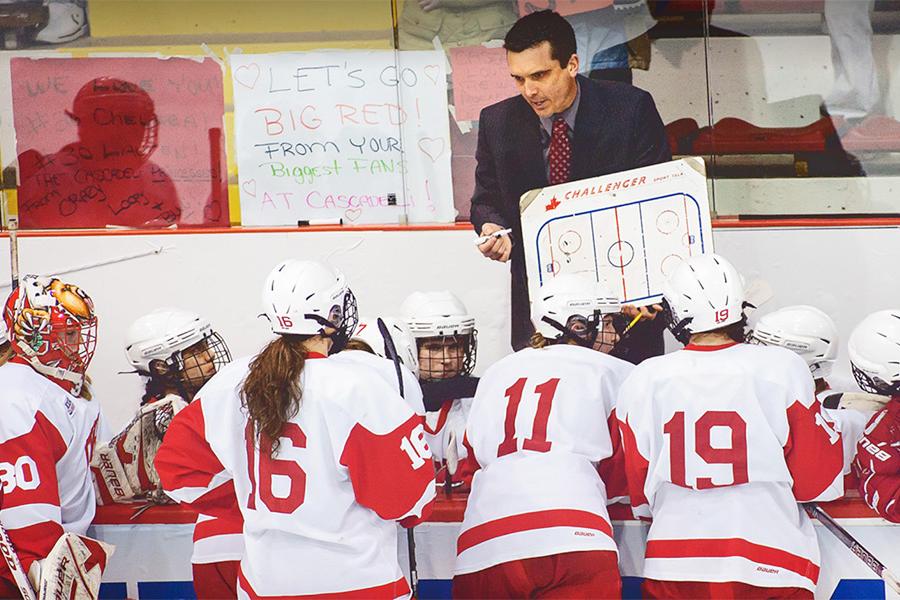 A coach with a white board in front of several hockey players in red and white jerseys