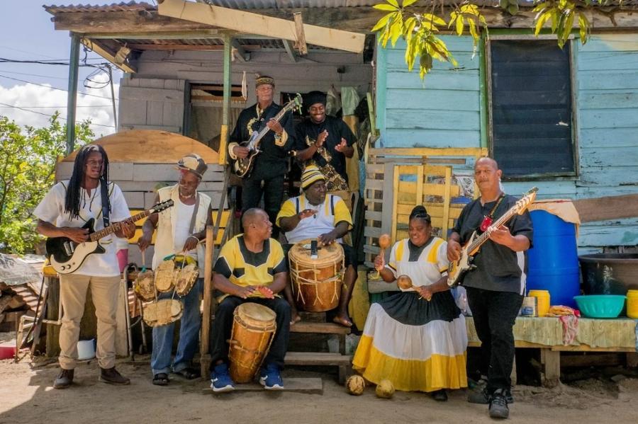 Several people standing around and sitting on porch steps in the shade, playing drums, guitars and maracas