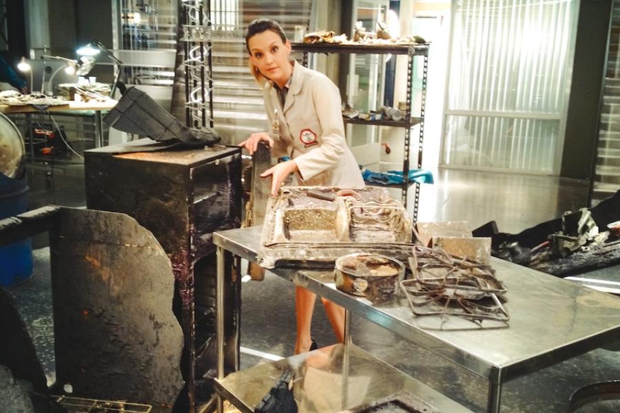 Actor on the set of a lab, wearing a chic trench coat, among a collection of burned or corrupted objects